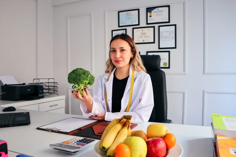 Female nutritionist in office holding broccoli, surrounded by fruits, promoting healthy lifestyle.